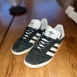 Adidas gazelle sneakers svart/vit - Säljer ett par klassiska Adidas gazelle sneakers i svart mocka med vita detaljer och de ikoniska tre ränderna på sidan. Skorna har snörning, vit platt sula med hexagonmönster undertill och rund tå. Tungan är vit med Adidas-logga. Perfekt för dig som gillar retrostil.