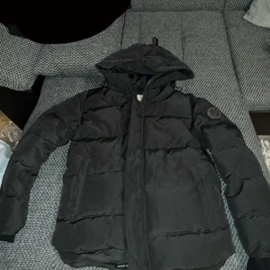 Svart pufferjacka Canada Goose M - Svart Canada Goose pufferjacka med huva och dragkedja framtill. Jackan har quiltad design och en rund Canada Goose-logga på vänster ärm. Perfekt för kalla dagar med sin tjocka och vadderade look. Passar dig som gillar streetwear och enkel stil. PRIS KAN DISKUTERAS!