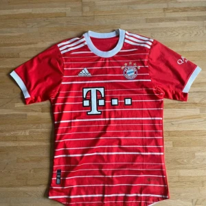 Röd Bayern München fotbollströja Adidas - Säljer en röd Bayern München matchtröja från Adidas med vita ränder och detaljer. Tröjan har klubbmärke, sponsortryck och korta ärmar med vit kant. Materialet är lätt och andas, perfekt för träning eller match. Snygg design med rund hals och tryck på ärmarna.