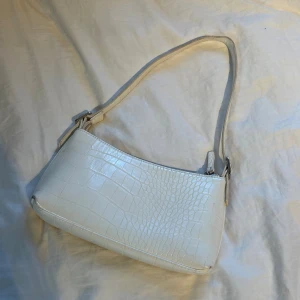 Vit axelväska i croco-imitation från NA-KD - Snygg vit axelväska från NA-KD med croco-imitation och stilren design. Väskan har justerbart axelband och stängs med dragkedja. Perfekt accessoar för att lyfta din outfit med en trendig touch.