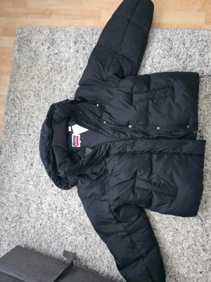 Svart pufferjacka från Levi's - Svart pufferjacka från Levi's med huva och dragkedja samt knappar framtill. jackan med dunfyllning (80 % dun, 20 % fjäder) som passar en person på 180 cm är sannolikt en varm och bekväm modell. Perfekt för kalla dagar.