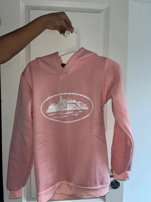Rosa hoodie från Rapidstreet - Snygg rosa hoodie från Rapidstreet med vit tryckt grafik på bröstet och en patch på ärmen. Tröjan har huva, långa ärmar och mjukt fleecefoder på insidan. Perfekt för dig som gillar streetwear och vill ha en chill look.