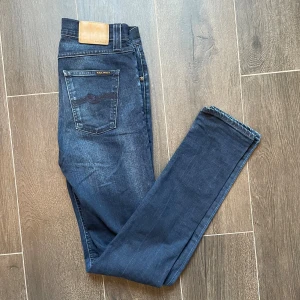 Nudie Jeans Mörkblå - Snygga mörkblå slim fit jeans från Nudie Jeans med klassisk femficksdesign och läderpatch bak. Jeansen har diskreta slitningar framtill och bak, samt stretchig känsla tack vare bomull och elastan. Perfekta för dig som gillar en smal passform och stilren look. För frågor eller prisdiskutering så är det bara att skriva😁