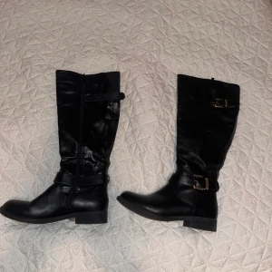 Svarta höga stövlar med spänne - Snygga svarta stövlar med platt sula och rund tå. Stövlarna har dekorativa spännen i metall och dragkedja på sidan för enkel på- och avtagning. Tillverkade i skinnimitation som ger en stilren look. Perfekta för dig som gillar klassisk och edgy stil.