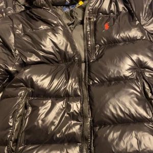 Svart pufferjacka från Polo Ralph Lauren - Svart pufferjacka från Polo Ralph Lauren med glansig finish och röd broderad logga på bröstet. Jackan har huva, dragkedja framtill och sidofickor med dragkedja. Perfekt för kalla dagar och har en riktigt schysst streetstil.