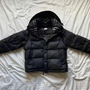 Svart Burberry pufferjacka med huva - Svart pufferjacka från Burberry med avtagbara ärmar, vilket gör att du kan bära den som väst. Jackan har en stor huva med klassiskt rutigt Burberry-foder i grått och vitt, samt dragkedja och knappar framtill. Två sidofickor med dragkedja och Burberry-logga på ärmen.