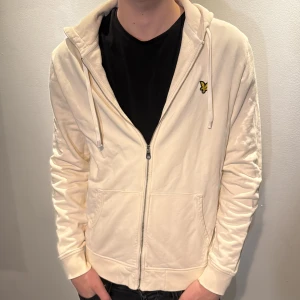 Beige hoodie från Lyle & Scott - Snygg beige hoodie från Lyle & Scott med dragkedja och klassisk gul logga på bröstet. Tröjan har huva med snörning, två fickor fram och är tillverkad i mjuk bomull. Perfekt för en chill och stilren look. Modellen är 186 cm och väger 70kg 