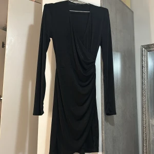 Svart omlottklänning från NLY One - Snygg svart kort omlottklänning från NLY One med lång ärm och v-ringning. Klänningen har draperad effekt framtill och är tillverkad i ett glansigt, stretchigt material som ger en elegant look.