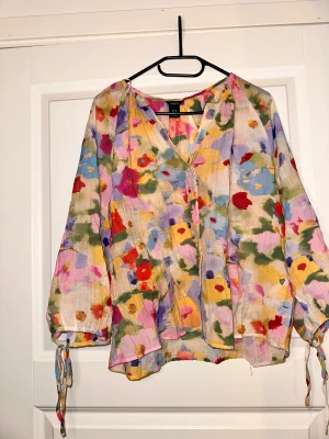 Blommig blus från Lindex i M - Färgglad blus från Lindex med blommigt mönster i rosa, gul, lila, röd, blå och grön. Luftig modell med v-ringning, långa ärmar och knytband vid ärmsluten. Perfekt för dig som gillar en bohemisk och lekfull stil.