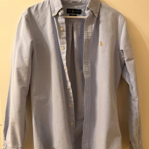 Ljusblå skjorta från Ralph Lauren i storlek M - Säljer en ljusblå skjorta från Ralph Lauren köpt på NK i slim fit med klassisk button down-krage och broderad logga på bröstet. Skjortan är långärmad och tillverkad i mjuk bomull. Perfekt för dig som gillar stilren och tidlös design. Passar bra om du är över 180cm lång.