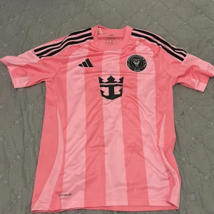 Inter Miami Messi #10 rosa fotbollströja - Säljer en officiell Inter Miami fotbollströja från Adidas i rosa med svarta detaljer och ränder på axlarna. Tröjan har korta ärmar, klubbmärke på bröstet och Messi #10 tryckt på ryggen. Tillverkad i lätt och ventilerande polyester med Aeroready-teknologi.