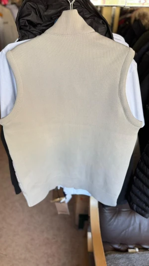 Beige dunväst från Burberry - Snygg beige dunväst från Burberry med ribbad krage och muddar. Västen har quiltad framsida, två dragkedjefickor och klassisk Burberry-logga på bröstet. Baksidan är helt ribbstickad och ger en clean look. Perfekt för lager-på-lager och stilrena outfits.