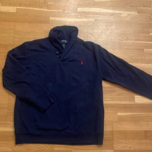Marinblå tröja från Polo Ralph Lauren XL - Snygg marinblå tröja från Polo Ralph Lauren i storlek XL. Tröjan har lång ärm, snörning vid halsen och den klassiska röda Polo-loggan broderad på bröstet. Materialet är mjuk bomull blandat med polyester för extra komfort.