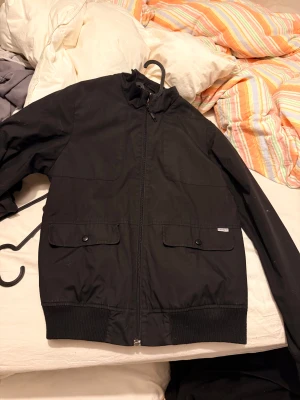 Svart  Carhartt Jacka - Svart bomberjacka från Carhartt med coolt mönstrat innerfoder i rött och vitt. Jackan har dragkedja framtill, två fickor med lock och knappar, samt ribbade muddar vid midja och ärmslut. Perfekt för dig som gillar streetwear och snygga detaljer.