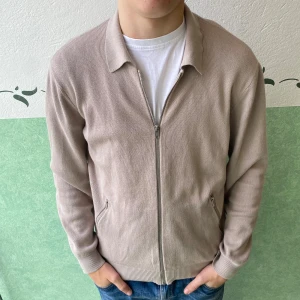 Beige stickad cardigan jacka från Riley - Snygg beige stickad cardigan jacka från Riley med dragkedja framtill och två fickor med dragkedja. Jackan har klassisk krage och ribbade muddar vid ärmslut och nederkant. Perfekt för dig som gillar stilren och enkel design.