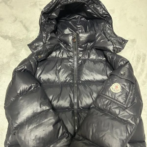 Svart Moncler pufferjacka med huva - Säljer en svart pufferjacka från Moncler med ikonisk logga på ärmen och avtagbar huva. Jackan har quiltad design, dragkedja framtill och praktiska fickor. Materialet är glansigt och tillverkat i polyester med dunfyllning, perfekt för kalla dagar.