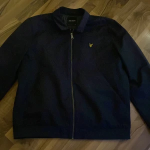 Mörkblå bomberjacka från Lyle & Scott - Snygg mörkblå bomberjacka från Lyle & Scott med klassisk gul logga på bröstet. Jackan har dragkedja framtill, krage och långa ärmar. Perfekt för dig som vill ha en stilren och enkel jacka med modern touch.