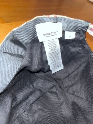 Burberry rutig keps i bomull - Snygg Burberry keps med klassiskt rutigt mönster i beige, svart och rött. Justerbar rem baktill med metallspänne. Tillverkad i bomull och har den ikoniska Burberry-looken som lyfter din stil till nästa nivå.