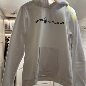 Vit hoodie från Sail Racing - Vit hoodie från Sail Racing med svart logotyptryck på bröstet. Tröjan har huva, magficka och långa ärmar. Mjuk insida och klassisk passform, perfekt för chill dagar. Snygg och enkel design som funkar till jeans eller mjukis.