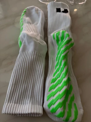 Vita träningsstrumpor med gröna ränder skill socks ej använda storlek-43-46 - Säljer ett par vita träningsstrumpor från Unisport med gröna zigzag-ränder undertill för extra grepp. Strumporna har ribbad design och svart logga upptill. Perfekta för fotboll eller gymmet, riktigt sköna och sitter bra på foten.