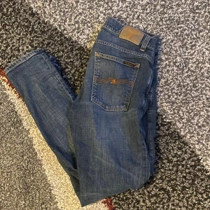Blå jeans från Nudie Jeans, storlek 33/32 - Snygga blå jeans från Nudie Jeans med klassisk orange söm på bakfickorna och logotyp på läderpatchen. Modellen har en smal passform och raka ben, tillverkade i slitstarkt denim med coola slitningar och detaljer. Perfekt för dig som gillar street och casual stil.