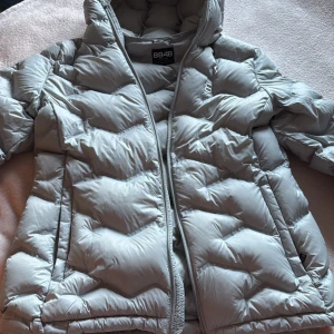 Ljusgrå pufferjacka från 8848 Altitude - Snygg ljusgrå pufferjacka från 8848 Altitude med huva och hel dragkedja. Jackan har ett quiltat mönster och är tillverkad i syntetmaterial. Perfekt för kalla dagar och har en modern, loose passform som ger en cool vibe.