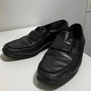 Svarta loafers med spänne i skinn - Säljer ett par svarta loafers från ECCO, tillverkade i Portugal. Skorna har en stilren design med rund tå, platt sula och ett dekorativt metallspänne över vristen. Materialet är mjukt skinn och de har en klassisk, tidlös look som passar till många outfits.