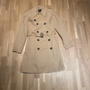 Beige trenchcoat från Lindex, strl 38 - Snygg beige trenchcoat från Lindex i klassisk modell med dubbelknäppning, bälte i midjan och stora kragar. Jackan har långa ärmar och svarta knappar framtill. Perfekt för dig som gillar stilrena och tidlösa plagg.