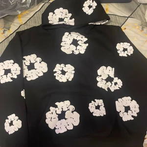 Svart hoodie från Denim Tears med blommor - Svart hoodie från Denim Tears med stora vita blommönster över hela tröjan. Klassisk huva och känguruficka framtill. Snygg och unik streetwear-stil som sticker ut i mängden.
