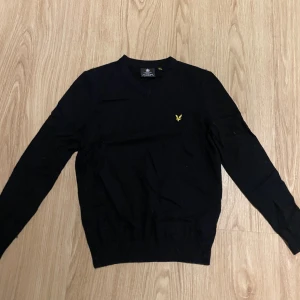 Svart ulltröja Lyle & Scott - Svart tröja från Lyle & Scott med gul broderad logga på bröstet. Tröjan har lång ärm och är tillverkad i mjuk ull och bomull. Perfekt för dig som gillar stilrena och klassiska plagg.