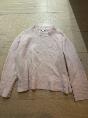 Ljusrosa stickad tröja med ribbade detaljer - Mysig ljusrosa stickad tröja med rund hals och breda ribbade muddar vid ärmslut och nederkant. Tröjan har en loose passform och är perfekt för kyliga dagar. Enkel och stilren design som är lätt att matcha med jeans eller kjol.