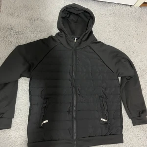 Svart cardigan  - Svart pufferjacka med huva och hel dragkedja framtill. Jackan har quiltad front och rygg, två sidofickor med dragkedja och mjuka muddar vid ärmslut och nederkant. Perfekt för dig som gillar en clean och sportig look.