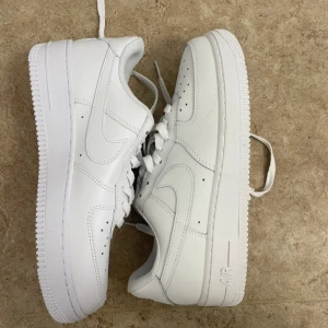 Nike Air Force 1 vita sneakers - Fräscha Nike Air Force 1 sneakers i helvitt skinn med klassisk rund tå och platt sula. Snygga detaljer som perforeringar på tån och tydlig Nike-logga på hälen. Lågt skaft och snörning framtill för en clean och tidlös look.