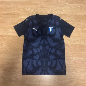Malmö FF x Puma blå fotbollströja - Snygg blå fotbollströja från Puma med Malmö FF-emblem på bröstet och vita Puma-loggor på axlarna. Tröjan har korta ärmar och ett mörkblått mönster framtill. Tillverkad i lätt och ventilerande material, perfekt för träning eller match.