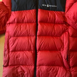 Röd och svart Sail Racing dunjacka - Säljer en fet dunjacka från Sail Racing i rött med svart axelparti och huva. Jackan har puffad design, dragkedja framtill och logga på bröstet. Perfekt för kalla dagar med sin tjocka och isolerande fyllning. Snygg streetstil och riktigt skön passform.
