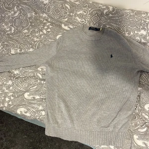 Grå stickad tröja Polo Ralph Lauren - Snygg grå stickad tröja från Polo Ralph Lauren i storlek M. Klassisk rund hals och långärmad modell med broderad marinblå logga på bröstet. Ribbstickade muddar vid ärmslut och nederkant ger en clean look. Perfekt för dig som gillar stilrena vibes.