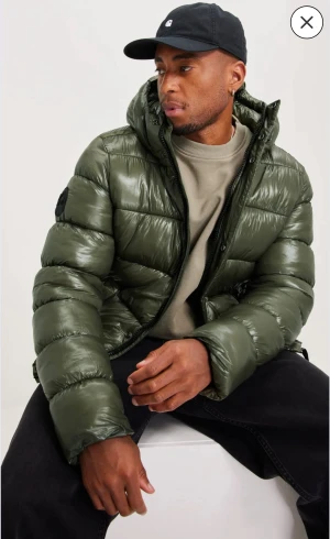 Grön pufferjacka från Superdry - Säljer en grön pufferjacka från Superdry med huva och glansig finish. Jackan har breda, quiltade sektioner och dragkedja framtill. Perfekt för dig som gillar streetwear och vill ha en varm jacka med cool look. Passar till både hoodie och t-shirt.