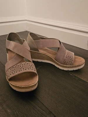 Beige kilklackade sandaler från Rieker - Snygga sandaler med kilklack från Rieker i beige mocka med blommigt perforerat mönster framtill. Skorna har korslagda remmar, öppen tå och en flätad detalj runt klacken. Perfekt för sommarens alla tillfällen och riktigt bekväma att gå i. Går såklart att diskutera priset💕🎀