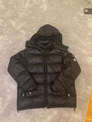 Svart Moncler pufferjacka med huva - Svart pufferjacka från Moncler med huva, dragkedja och fickor med detaljerade knappar. Jackan har Moncler-logga på ärmen och quiltad design. Materialet är glansig polyester och dunfyllning, perfekt för kalla dagar. Snygg och klassisk look med långärmad passform.