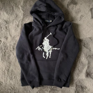 Svart hoodie från Polo Ralph Lauren - Svart hoodie från Polo Ralph Lauren med stor vit logga och polospelare på bröstet. Tröjan har huva med snörning och ribbade muddar vid ärmslut och nederkant. Tillverkad i mjuk bomull, perfekt för chill dagar. Klassisk och stilren design.