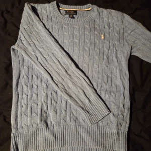 Ljusblå stickad tröja Polo Ralph Lauren - Snygg ljusblå stickad tröja från Polo Ralph Lauren med klassiskt kabelstickat mönster och rund hals. Tröjan har långa ärmar, ribbade muddar och en liten broderad logga i vitt på bröstet.