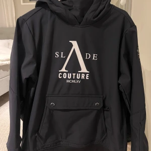 Skidjacka - Svart skidjacka från Slade Couture med stor vit logga framtill och mindre tryck baktill. Tröjan har huva, lång ärm och en stor ficka med knappar på magen. Materialet känns mjukt och har en lite loose passform.                                            Nypris 999kr