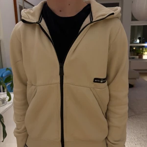 Beige hoodie från Sail Racing - Snygg beige hoodie från Snil Racing med svart dragkedja och huva. Tröjan har ett svart logomärke på bröstet och coolt tryck på ärmen. Tillverkad i mjuk bomull, perfekt för chill dagar. Fickor framtill och stilren design.