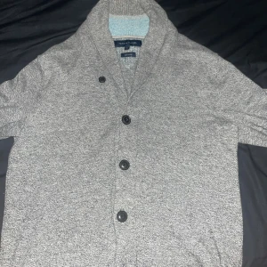 Grå kofta från Tommy Hilfiger - Snygg gråmelerad kofta från Tommy Hilfiger i mjuk ull. Koftan har bred krage, tre svarta knappar framtill och en chill, klassisk look. Perfekt för lager-på-lager och passar dig som gillar stilren design.