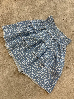 Blå blommig volangkjol med smock - Supersöt blå kortkjol med vitt blommönster och bred smock i midjan. Kjolen har flera volanger och är gjord i ett lätt, mjukt material som känns skönt mot huden. Perfekt för dig som gillar romantisk och feminin stil med extra volym.