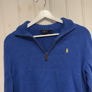 Blå stickad tröja Polo Ralph Lauren - Snygg blå stickad tröja från Polo Ralph Lauren i storlek XS. Tröjan har en ribbad krage, half zip-dragkedja och det klassiska gula Polo-logot broderat på bröstet. Perfekt för dig som gillar stilrena och tidlösa plagg. Hör av er med prisförslag och funderingar!
