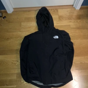 Svart vindjacka från The North Face - Svart vindjacka från The North Face med huva och reflekterande detalj vid fickan. Jackan har dragkedja framtill, elastiska muddar och meshfoder på insidan. Perfekt för blåsiga dagar och har en clean look med logga på bröstet.