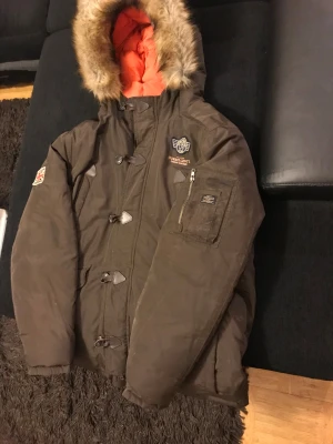 Brun parkajacka Morris Gents köp prise 10000 XL - Parkajacka från Morris Gents i brun färg med orange quiltat foder och äkta päls på huvan. Jackan har broderade loggor, patch på ärmen, dragkedja och knappar framtill samt praktiska fickor. Snygga detaljer som snörning och robust look.