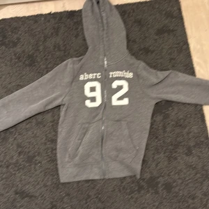 Grå hoodie Abercrombie Kids XL - Grå hoodie från Abercrombie Kids i storlek XL med vit tryckt text och siffran 92 på bröstet. Tröjan har huva, dragkedja och två fickor framtill. Mjuk insida och klassisk streetstil, perfekt för chill dagar. Snyggt och enkelt plagg som funkar året runt.