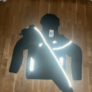 Svart Nike vindjacka med reflexdetaljer - Svart vindjacka från Nike med huva och hel dragkedja. Jackan har vita reflexdetaljer längs bröstet och vid fickorna, samt Nike-logga på bröstet. Perfekt för dig som vill sticka ut i mörkret och hålla stilen. Snygg och sportig design med praktiska fickor.